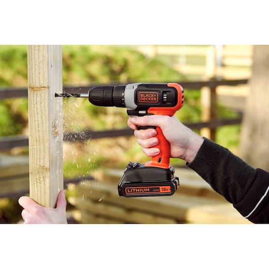 BLACK+DECKER BCD001C1 cordless drill with orange and black body, drilling into wood outdoors.