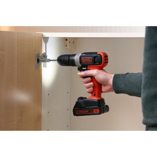 BLACK+DECKER BCD001C2K cordless drill in use installing a cabinet hinge, side view, orange and black color.