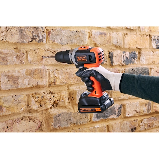 BLACK+DECKER BCD003C2K cordless drill in use on a brick wall, orange and black design, gloved hand holding tool.