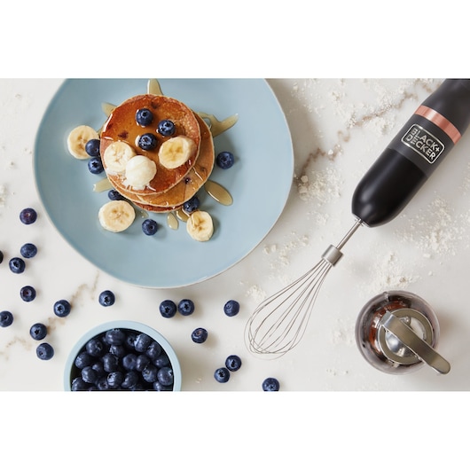 Photo of BLACK+DECKER BCKM1011KGF hand mixer with whisk attachment on white surface, pancakes, fruit, and syrup visible.