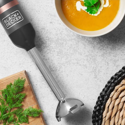 BLACK+DECKER BCKM1016KSG hand blender with stainless steel shaft on kitchen counter near soup bowl and herbs.
