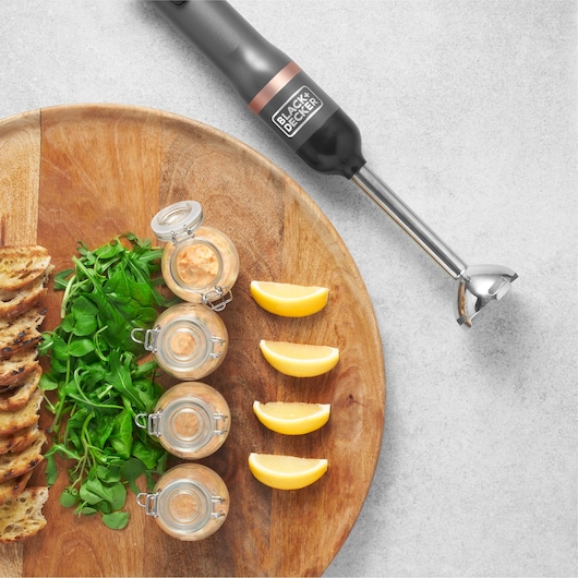 Photo of BLACK+DECKER BCKM1011KGF hand blender with food, wooden board, greens, toast, jars, and lemon wedges.
