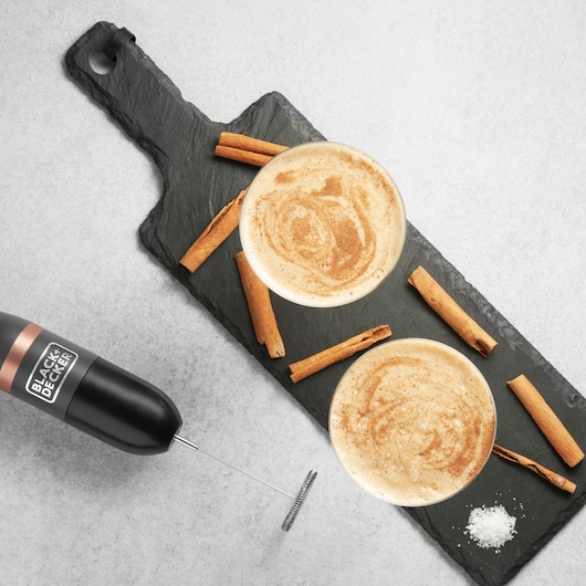 BLACK+DECKER BCKM1011KGF milk frother beside two cinnamon-topped lattes and cinnamon sticks on slate board.
