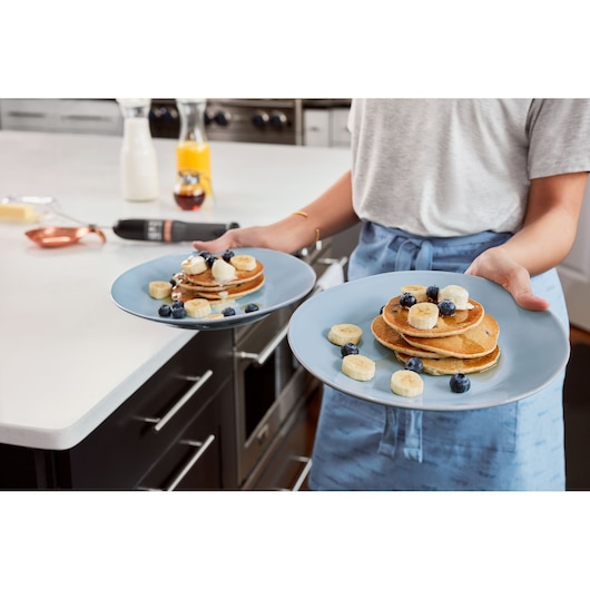 BLACKDECKER BCKM1011KGF photo of person holding two blue plates with pancakes, banana slices, and blueberries in kitchen.