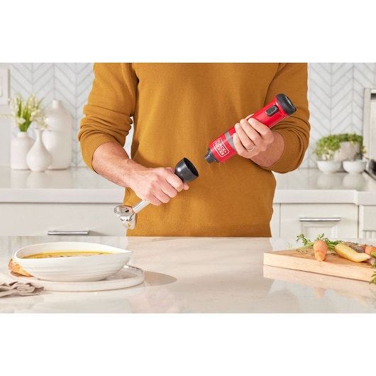 BLACK+DECKER BCKM1012KR cordless red hand blender being assembled in a kitchen near a bowl of soup and vegetables.
