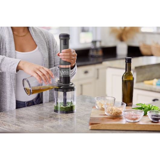 Photo of BLACK+DECKER BCKM101FP hand blender blending herbs on kitchen counter with ingredients and oil nearby.