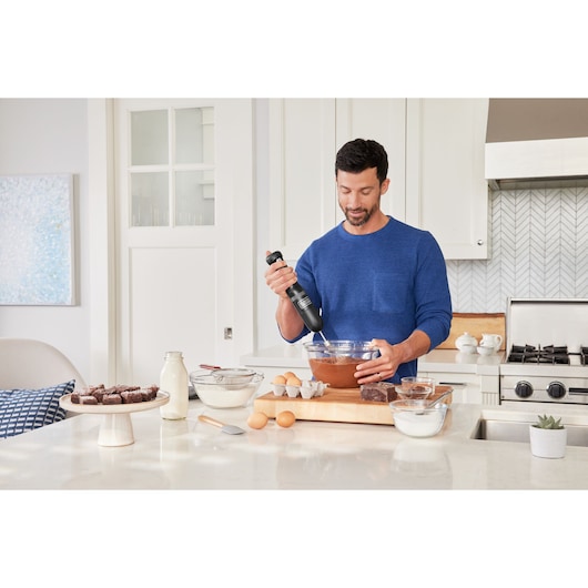 Photo of a person using a BLACKDECKER BCKM101WHFF hand mixer in a modern kitchen with baking ingredients.