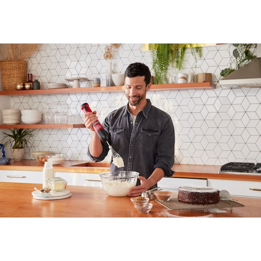Photo of a person using a BLACKDECKER BCKM101WHFF hand mixer to whisk ingredients in a glass bowl on a kitchen counter.