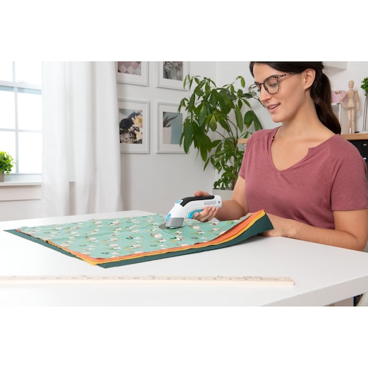 BLACKDECKER BCSC115 handheld fabric cutter in use on layered floral cloth in a home workspace.