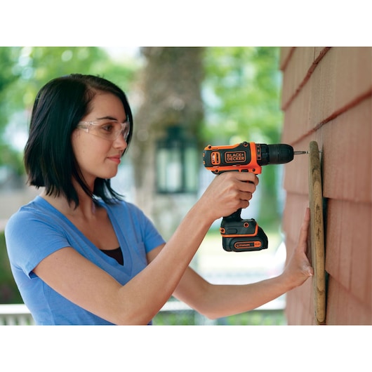 BLACKDECKER BDCDD12K cordless drill in use, orange and black, outdoors, drilling into wood siding.