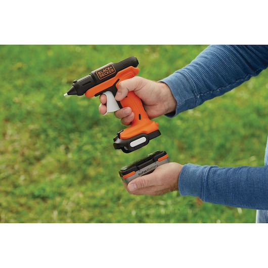 Photo of BLACK+DECKER BDCGG12N cordless glue gun with orange and black design, person attaching GoPak battery outdoors.