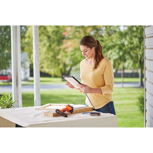 BLACKDECKER BDCGG12N cordless glue gun on outdoor table with wood pieces and blue glue stick, person using tablet nearby.