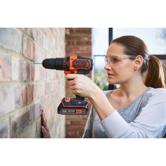Photo of BLACK+DECKER BDCHD18K cordless drill in use drilling a brick wall, orange and black design.