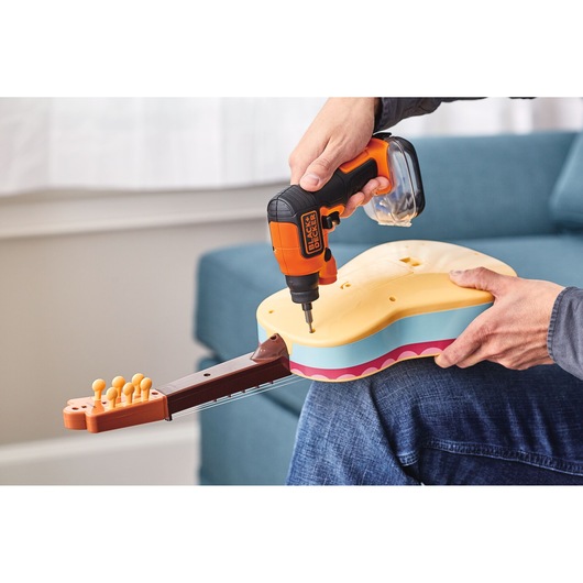 A person is using a BLACK+DECKER cordless screwdriver to assemble a colorful toy guitar, with the instrument resting on their lap.