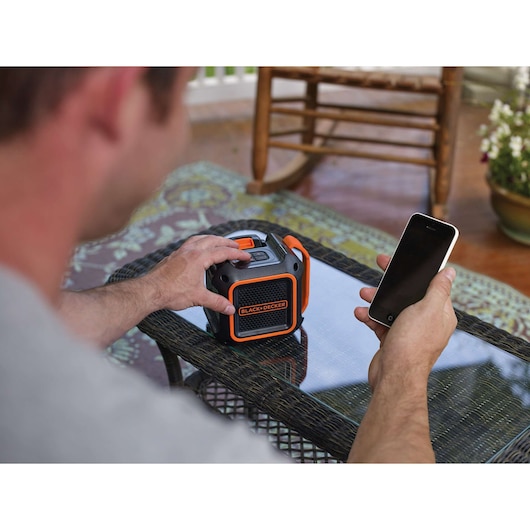 BLACK+DECKER BDCSP18N portable speaker with orange accents on glass table, person holding smartphone.