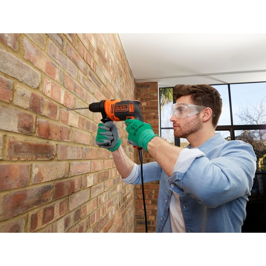 Person using BLACK+DECKER BEH710 corded drill on brick wall indoors, wearing gloves.