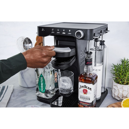 BLACKDECKER BEHB101 cocktail maker front angle with hand inserting pod, surrounded by liquor bottles and glass with ice.