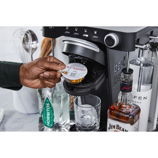 BLACK+DECKER BEHB101 beverage maker with hand inserting cocktail pod, liquor bottles and glass with ice nearby, front angle.