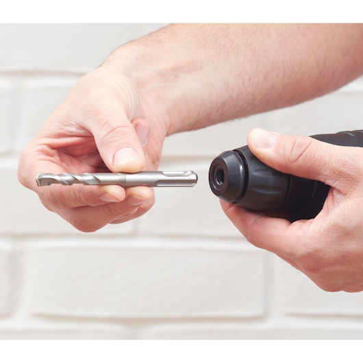 Photo of hands holding a BLACKDECKER BEHS01K drill and a metal drill bit near a white brick wall.