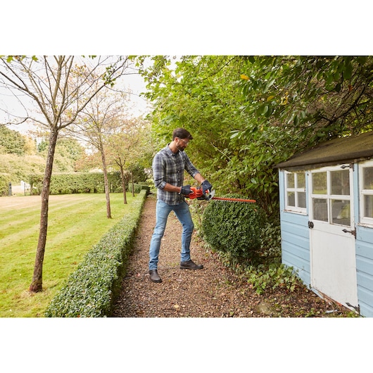 BLACKDECKER BEHT251 hedge trimmer in use trimming bushes in a garden near a blue shed.