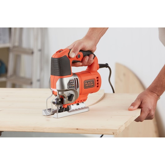 A person is using a BLACK+DECKER BES610 electric jigsaw to cut a piece of wood. The orange and black jigsaw is plugged in, and the hand grips the tool firmly. The scene includes woodworking materials and tools in the background.