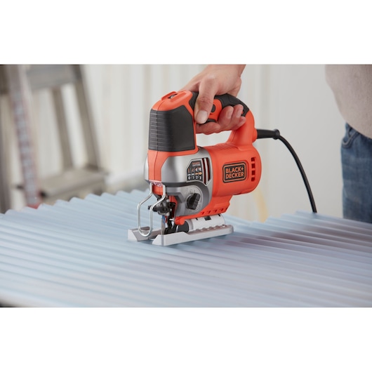 A person using a BLACK+DECKER BES610 electric jigsaw to cut a corrugated metal sheet. The jigsaw is orange and black, held firmly by hand, and plugged in for use.