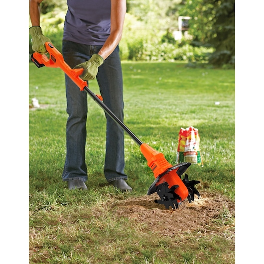 BLACK+DECKER BETL1820L orange cordless garden tiller in use on lawn, user wearing gloves outdoors.