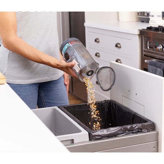 BLACKDECKER BHFEV36B2DW vacuum dust container being emptied into kitchen trash bin, angled side view.