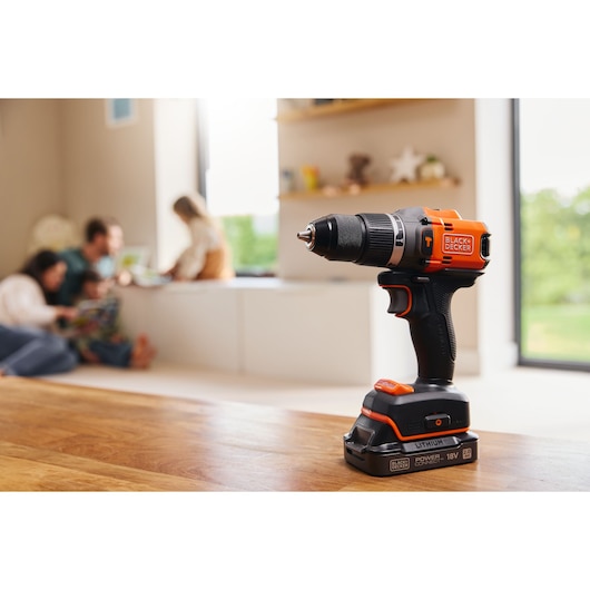 Photo of BLACK+DECKER cordless drill BLD683D1 with black and orange design on wooden table, blurred family in background.