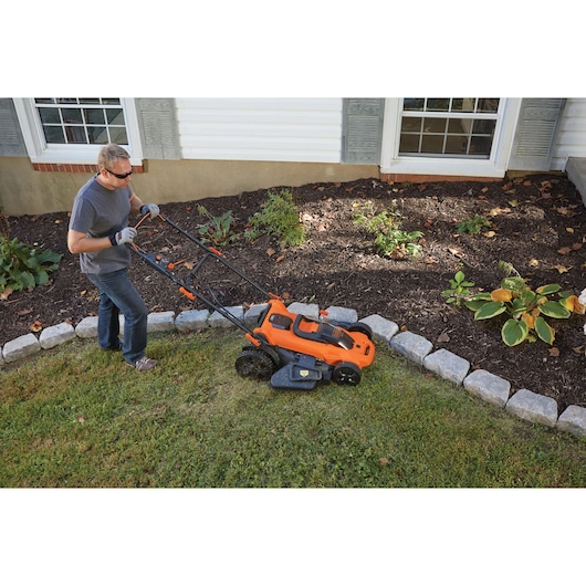 BLACKDECKER CLMA4825L2 orange cordless lawn mower on grass near garden bed, person pushing.
