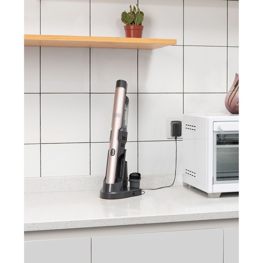 BLACKDECKER DVC320BRG handheld vacuum in rose gold charging on a kitchen counter beside a white oven.