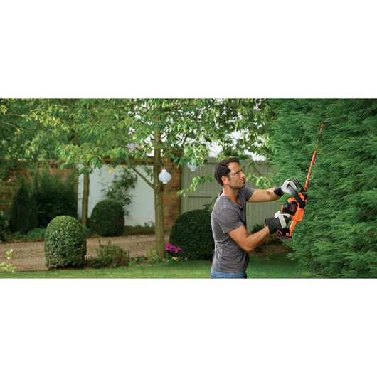 Photo of person using BLACK+DECKER GTC18452PCB cordless hedge trimmer to cut green hedge in garden.