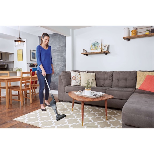 A person using a BLACK+DECKER stick vacuum cleaner to clean a patterned rug in a modern living room. The room features a gray sectional sofa, a wooden coffee table with a plant in a pot, and shelves with decorative items on the wall.