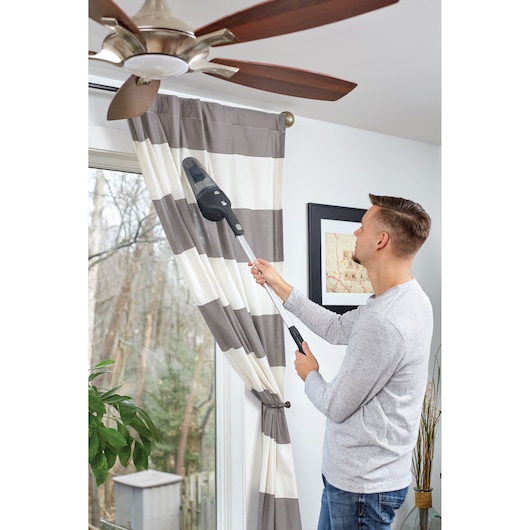 A person using a BLACK+DECKER NSVA315J cordless handheld vacuum cleaner with an extension to clean the top of window curtains in a home. The vacuum cleaner is directed toward dust on the curtains near a ceiling fan.