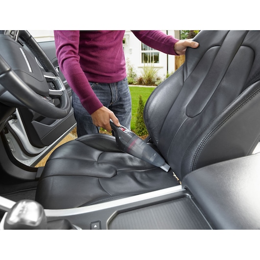 A person is using a BLACK+DECKER handheld vacuum cleaner to tidy up crumbs from the driver seat of a car with black leather upholstery.
