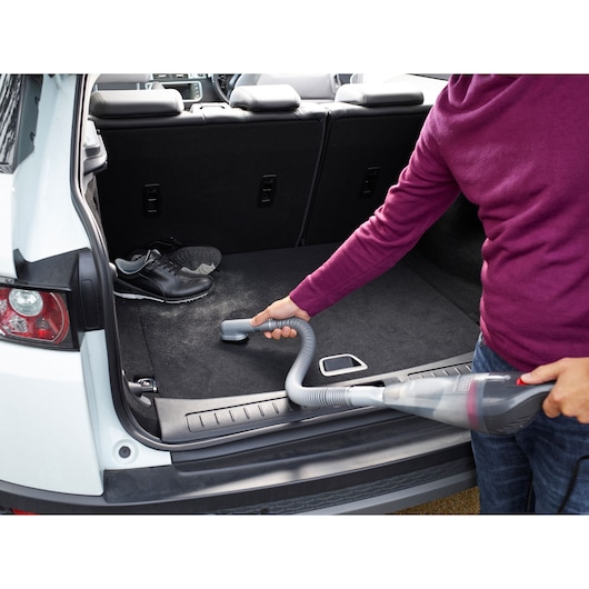 A person uses a BLACK+DECKER handheld vacuum cleaner with a flexible hose to clean the trunk of a car. There is a pair of black sneakers inside the car trunk.