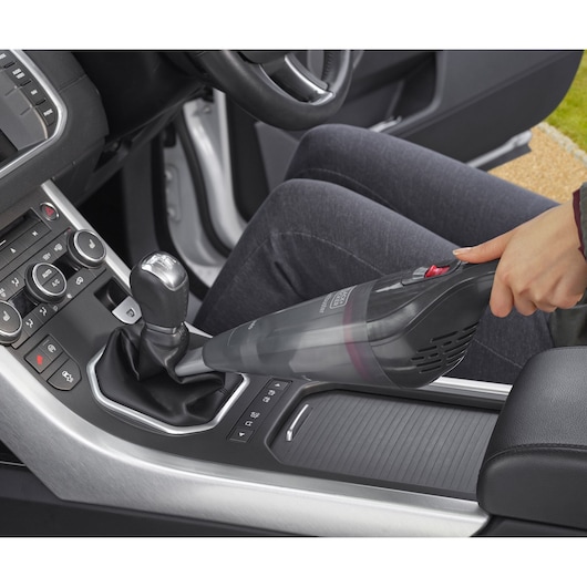 A person uses a BLACK+DECKER handheld vacuum cleaner to clean the interior between the gear shift and console of a car.