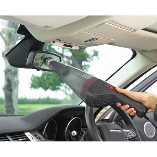 A person using a BLACK+DECKER handheld vacuum cleaner to clean the area around the rear-view mirror inside a car.