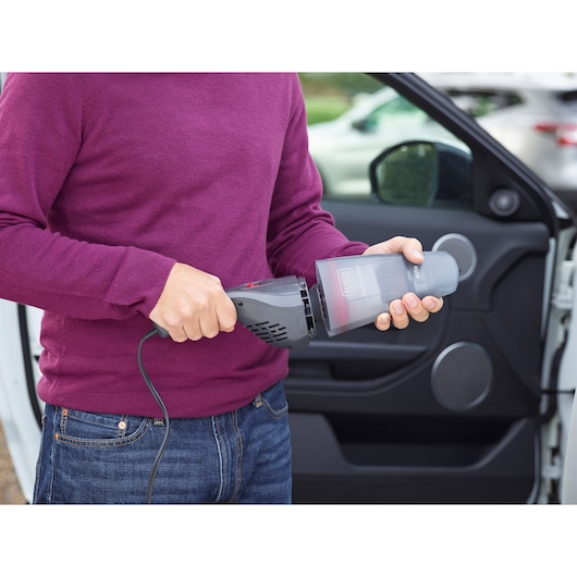 A person wearing a purple sweater is holding a BLACK+DECKER handheld vacuum cleaner NVB12AVA, standing next to an open car door.