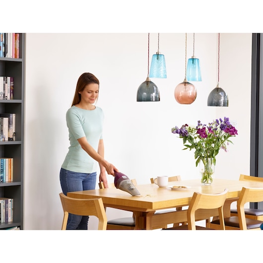 BLACKDECKER NVC115W handheld vacuum in use cleaning crumbs from a wooden table in a dining room setting.