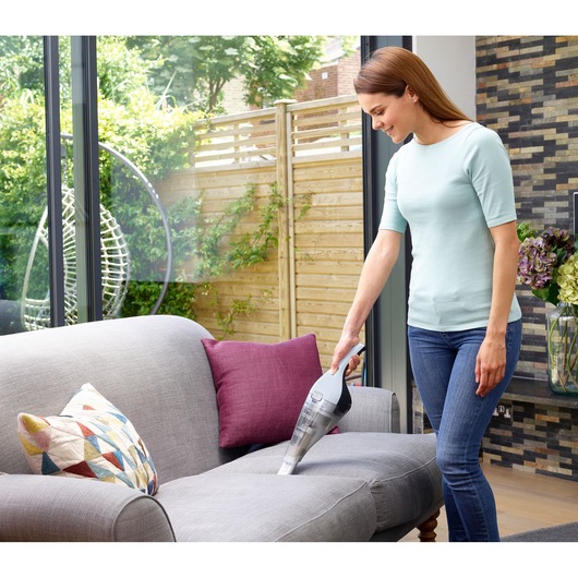 A person using a BLACK+DECKER handheld vacuum cleaner (model NVC215W) to clean a grey sofa in a living room with a modern design. The living room has large windows, colorful cushions, and a view of a wooden fence and greenery outside.