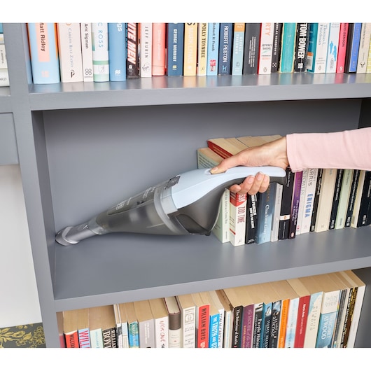 A hand using a BLACK+DECKER handheld vacuum cleaner to clean dust from a grey bookshelf filled with books.