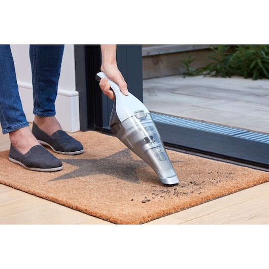 A person uses a BLACK+DECKER handheld vacuum cleaner to clean dirt from a doormat at a doorway.