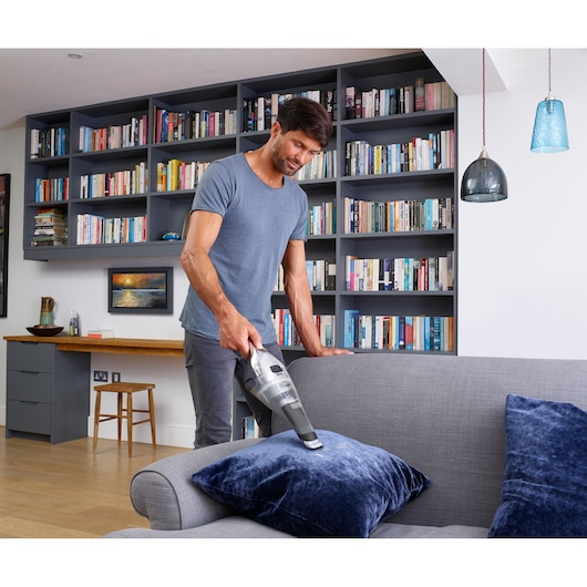 A person using a BLACK+DECKER handheld vacuum cleaner to clean a blue velvet cushion on a grey sofa in a modern living room with bookshelves in the background.