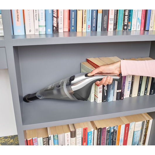 A person using a BLACK+DECKER handheld vacuum cleaner to clean the surface of a bookshelf with books.