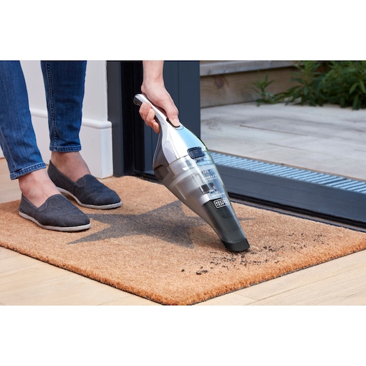A person is using a BLACK+DECKER handheld vacuum cleaner to remove dirt from a doormat near a doorway.