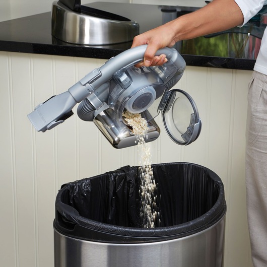 A person empties the contents of a BLACK+DECKER handheld vacuum cleaner into a trash bin with a black liner in a kitchen setting.