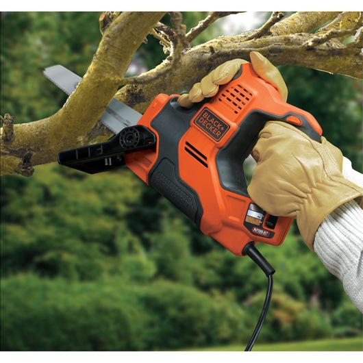 A person wearing gloves uses a BLACK+DECKER electric reciprocating saw to cut a tree branch outdoors.