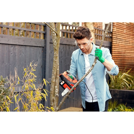 A person wearing green gloves and a light blue shirt is using a BLACK+DECKER electric saw to cut a tree branch in a garden with a wooden fence in the background.
