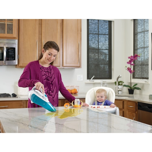 A person uses a BLACK+DECKER handheld vacuum cleaner (model WDA315J) to clean up a liquid spill on a kitchen countertop while a baby sits in a high chair nearby.